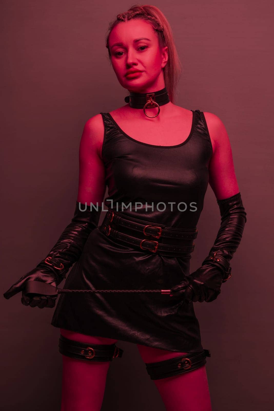 Beautiful dominant brunette mistress woman in latex dress and gloves posing with riding crop on red light backgound