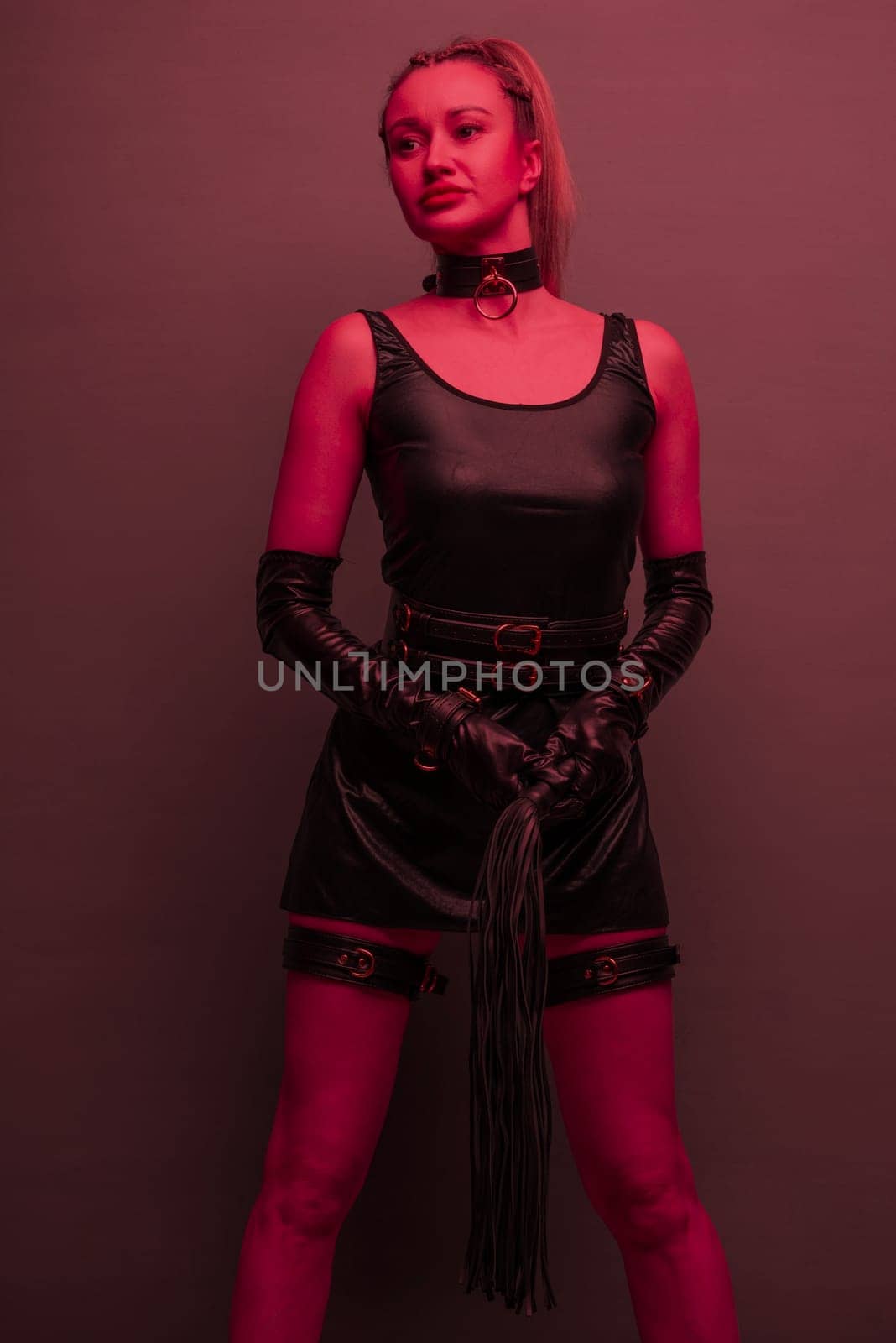 Beautiful dominant brunette mistress woman in latex dress and gloves posing with riding crop on red light backgound by zartarn