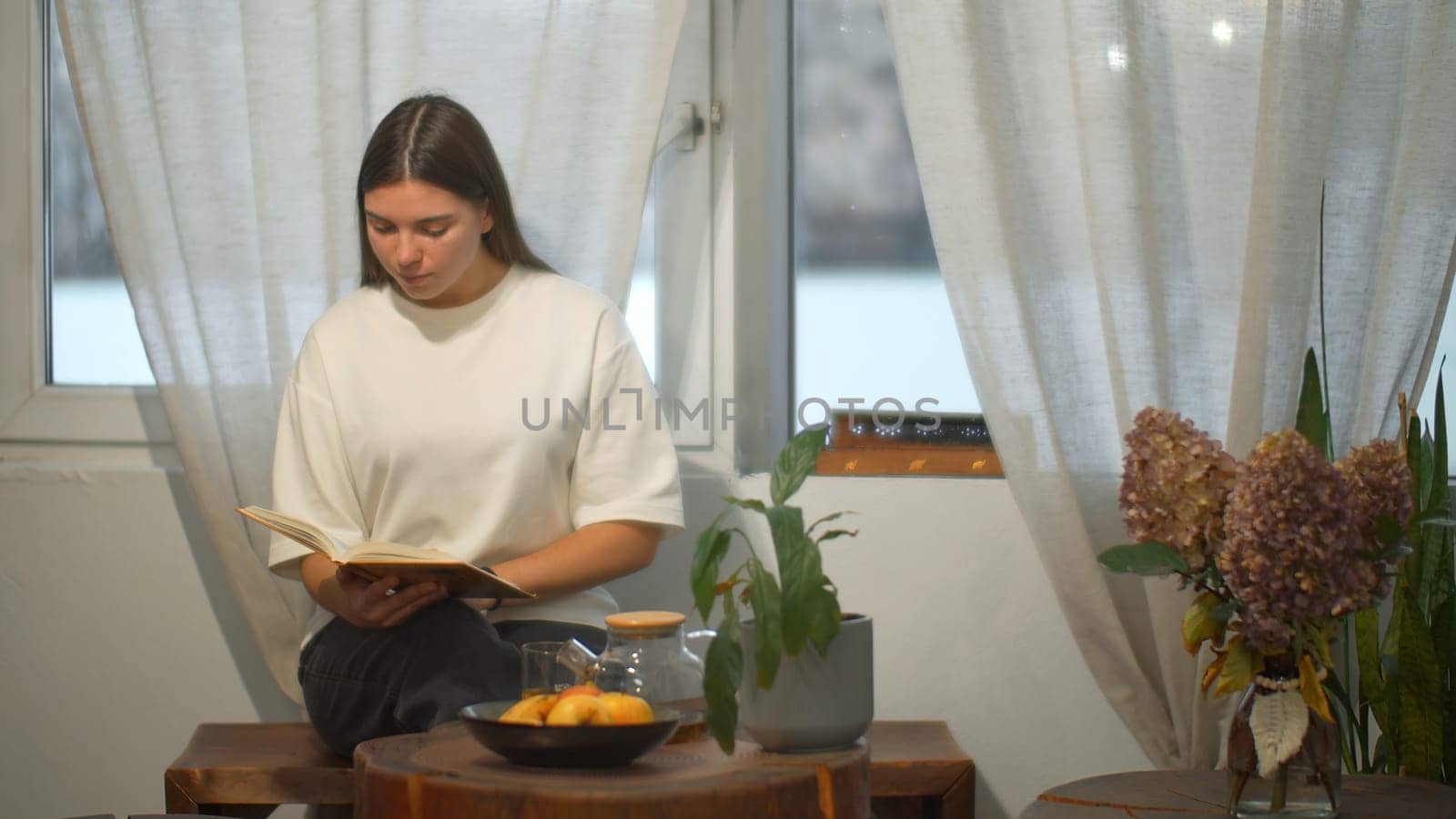 Young female student reads book in cafe. Media. Beautiful young woman comfortably spending time reading book in cafe. Cozy winter day in cafe with book and tea by Mediawhalestock