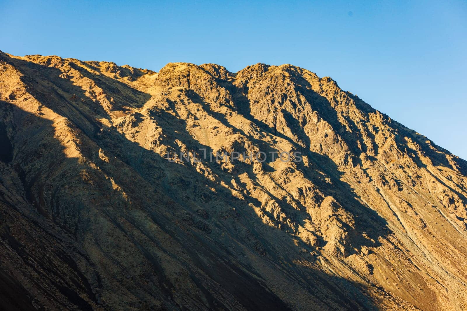 Natural landscape featuring a close up telephoto view of a mountain side slope under direct evening sun light with clear blue sky above.