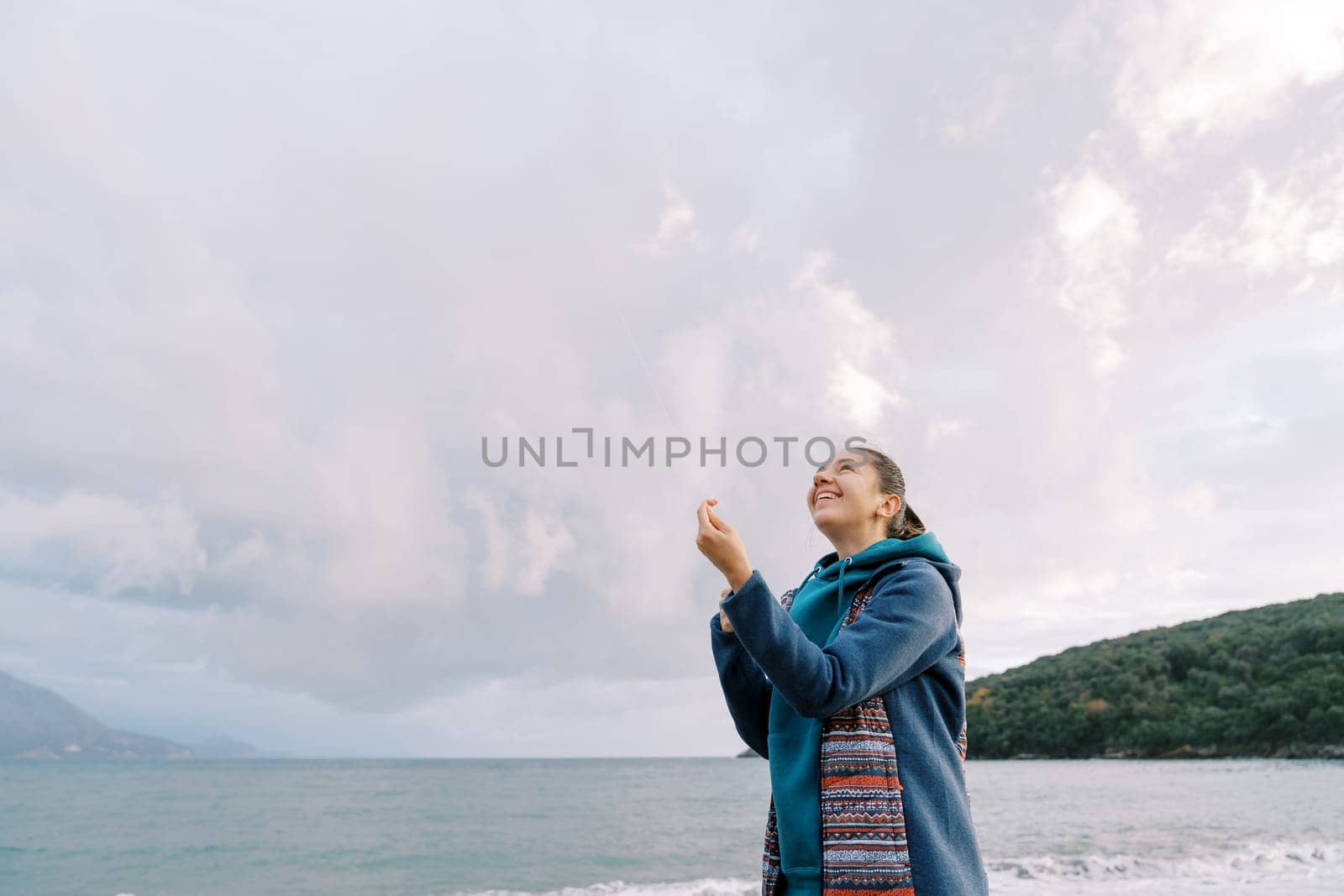 Young woman is standing by the sea with a kite spool in her hands, straightening the thread, and looking up. High quality photo