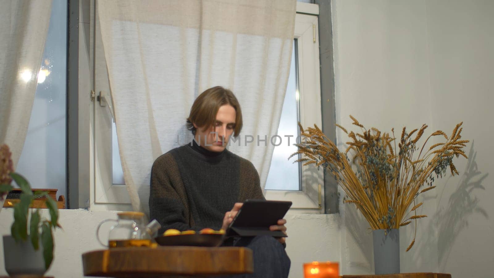 Young man drinks tea and works on tablet in cafe. Media. Student is relaxing with tablet and tea in cozy cafe. Handsome student studying on break in cafe by Mediawhalestock