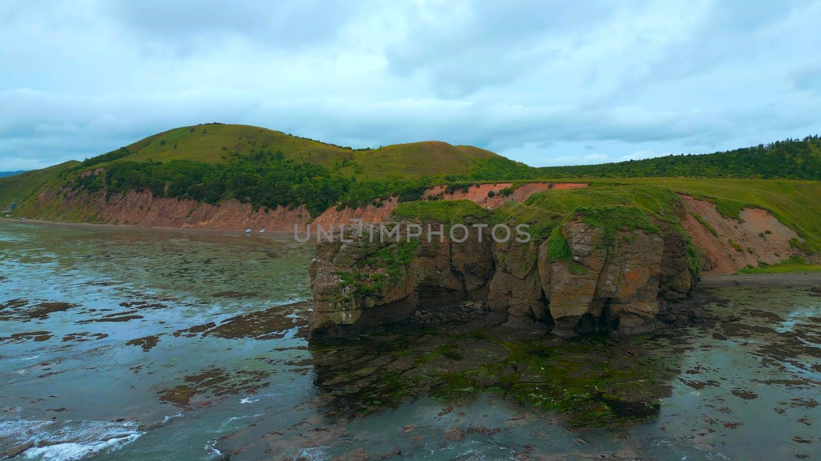 Top view of rocky coast with algae on shore. Clip. Landscape of rocks on coast with algae on background of mountains in cloudy weather. Diversity of flora of sea coast with rocky mountains.