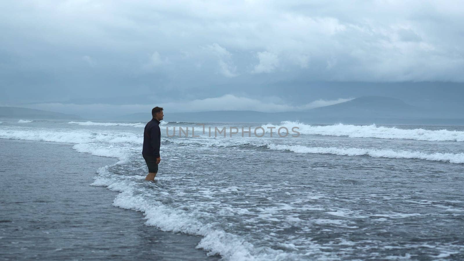 Man stands on water on seashore in cloudy weather. Clip. Lonely man stands in water with waves. Man stands in sea with waves on background of mountain coast.