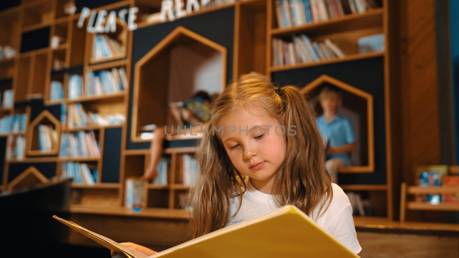 Young smart caucasian girl picking reading a book while sitting at library. Clever child learning, studying, open a books at library. Attractive kid turning page with blurring background. Erudition.