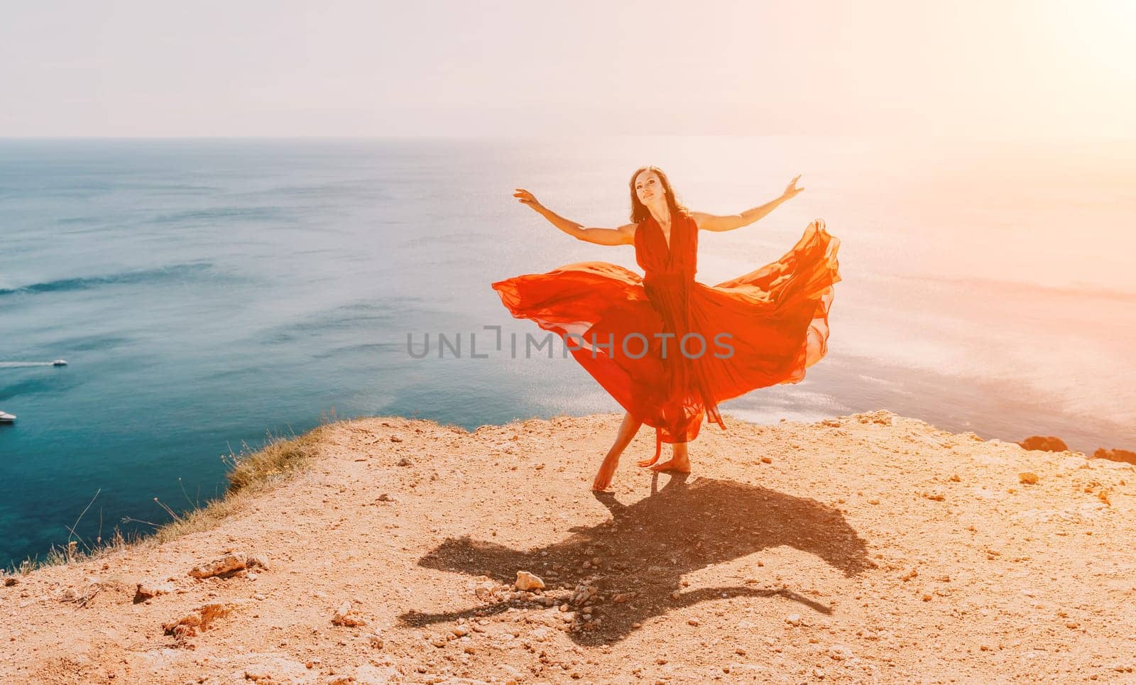 Side view a Young beautiful sensual woman in a red long dress posing on a rock high above the sea during sunrise. Girl on the nature on blue sky background. Fashion photo.