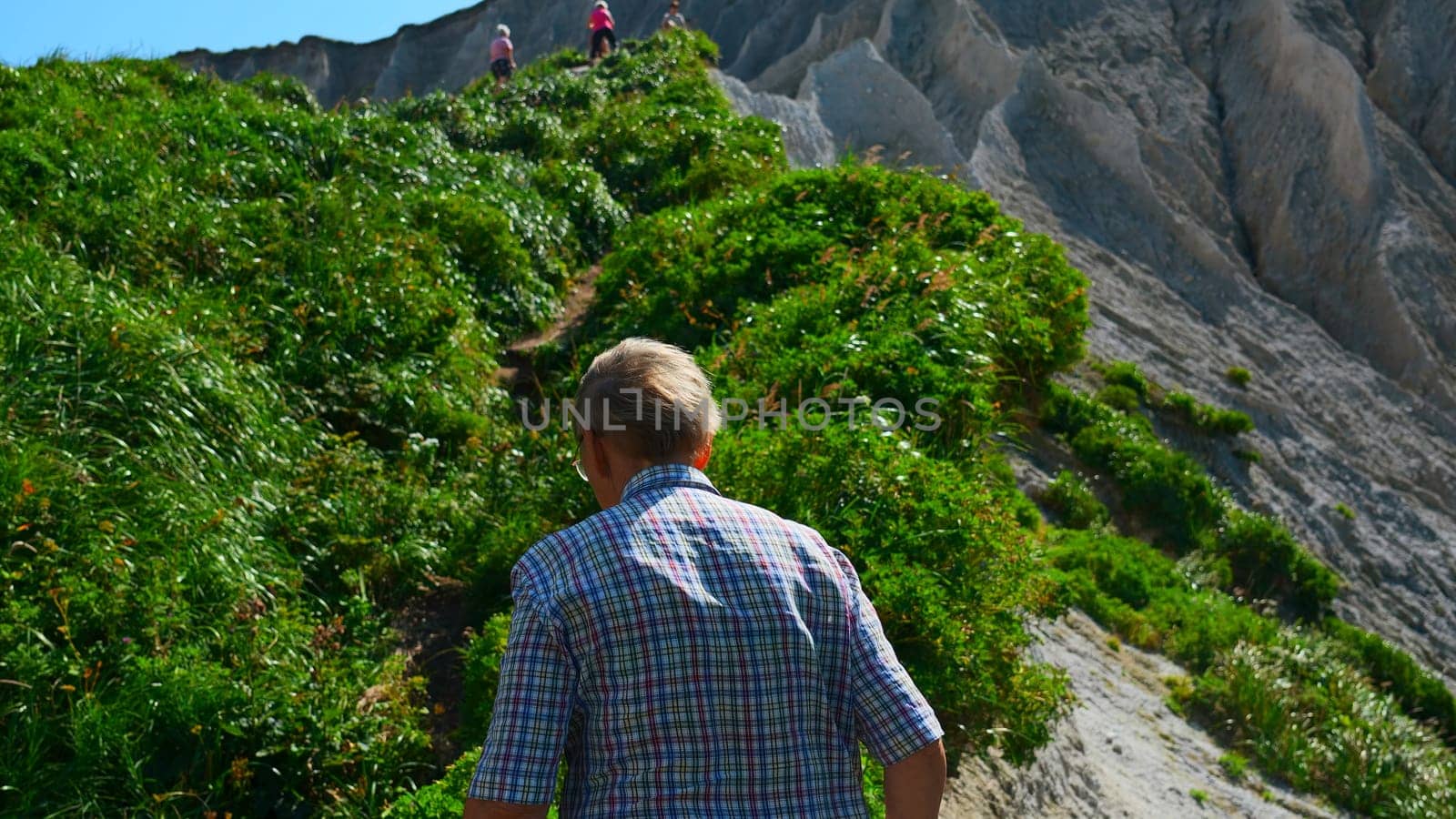 Grandfather on hike in mountains in summer. Clip. Elderly man walks along mountain path on sunny summer day. Rear view of elderly man walking on hike in hills with green grass by Mediawhalestock
