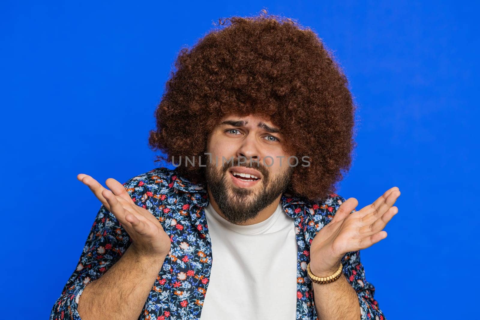 What. Why. Angry sad Caucasian man raising hands in indignant expression, asking reason of failure, demonstrating disbelief irritation by troubles. Stylish guy isolated on blue studio background