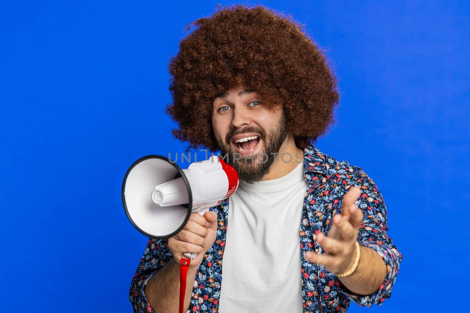 Caucasian man talking with megaphone, proclaiming news, loudly announcing advertisement discounts sale, using loudspeaker to shout speech. Happy stylish guy isolated on blue background. Copy-space