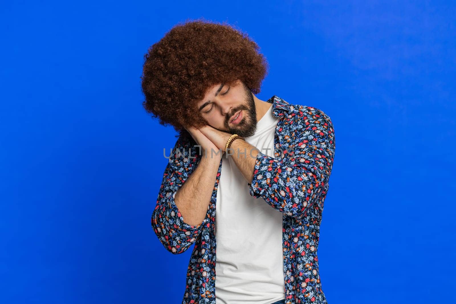 Tired exhausted young adult sleepy man with Afro hairstyle wig inattentive feeling somnolent lazy bored gaping suffering from lack of sleep, falling asleep. Guy isolated on studio blue background