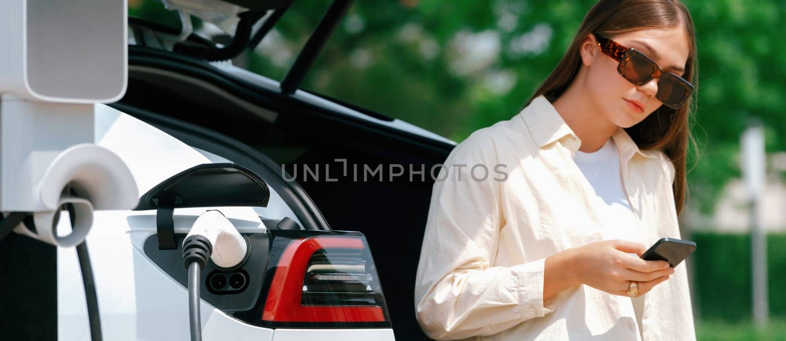 Young woman use smartphone to pay for electricity at public EV car charging station green city park. Modern environmental and sustainable urban lifestyle with EV vehicle. Panorama Expedient