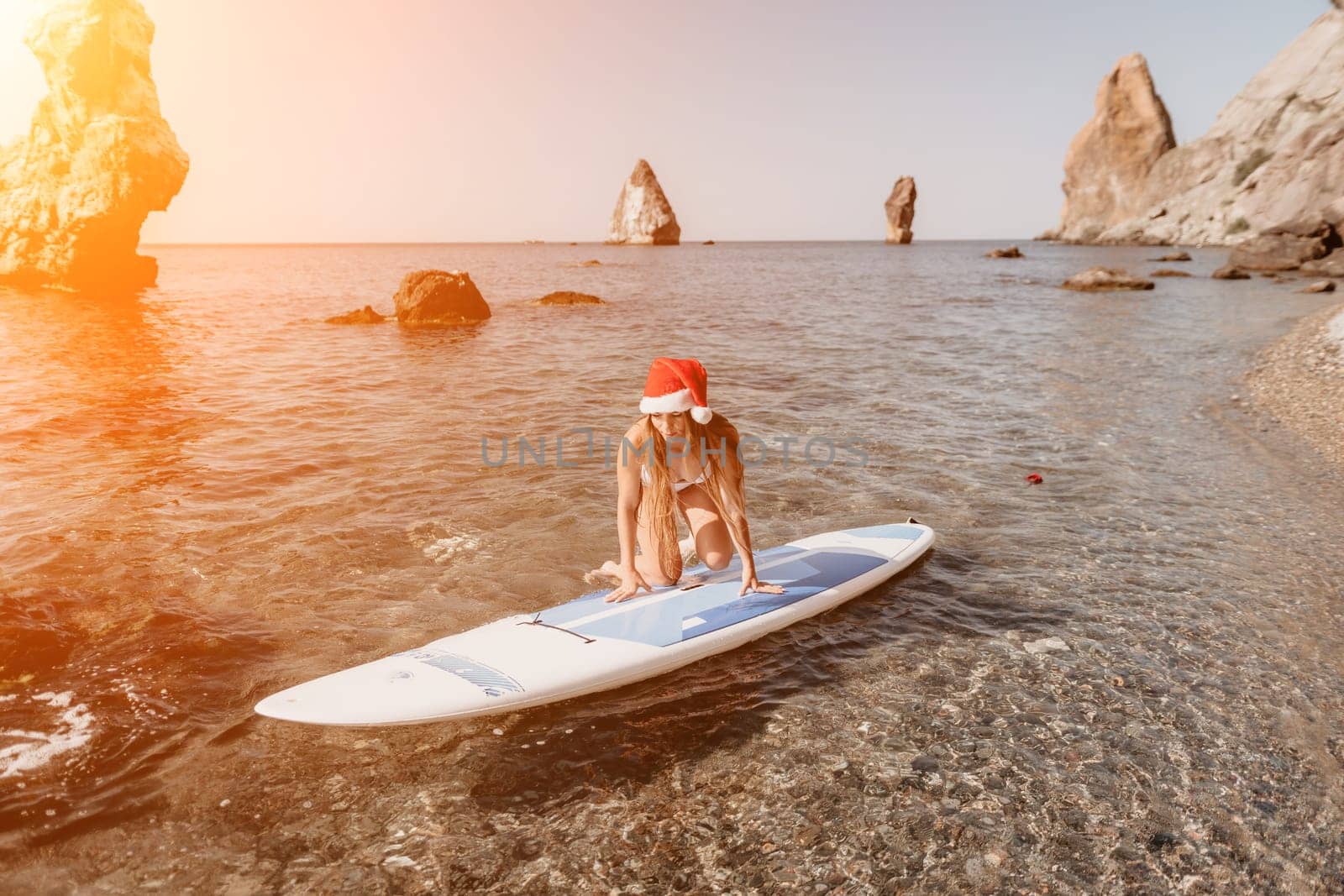 Woman sea sup. Close up portrait of happy young caucasian woman with long hair in Santa hat looking at camera and smiling. Cute woman portrait in a white bikini posing on sup board in the sea by panophotograph