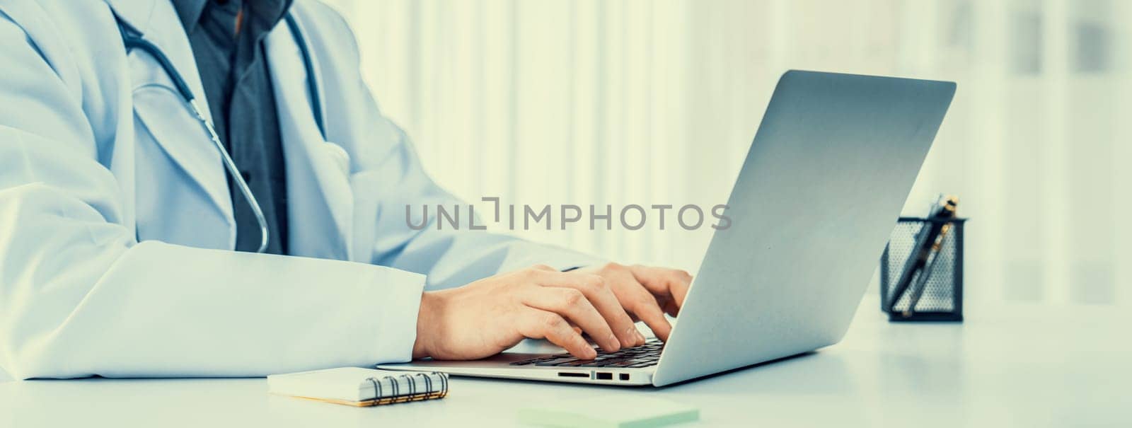 Doctor at hospital sit at his desk working on laptop diagnosing patient test results, developing treatment plan for illnesses and sicknesses. Medical staff and healthcare service. Panorama Rigid