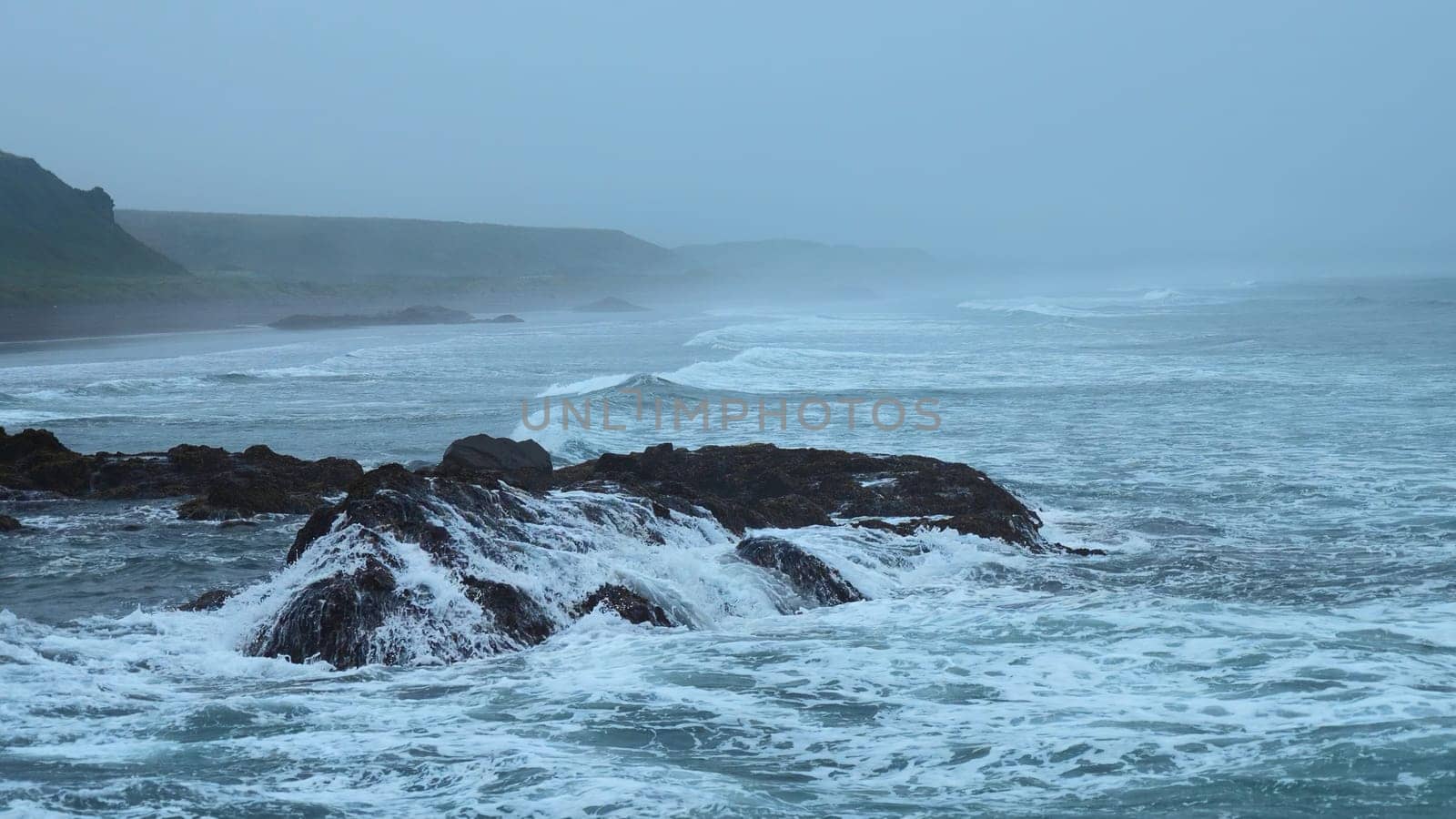 Beautiful waves break on rocks of rocky shore. Clip. Stone shore with storm waves in cloudy weather. Dramatic landscape of stone coast with beautiful splashes of waves.