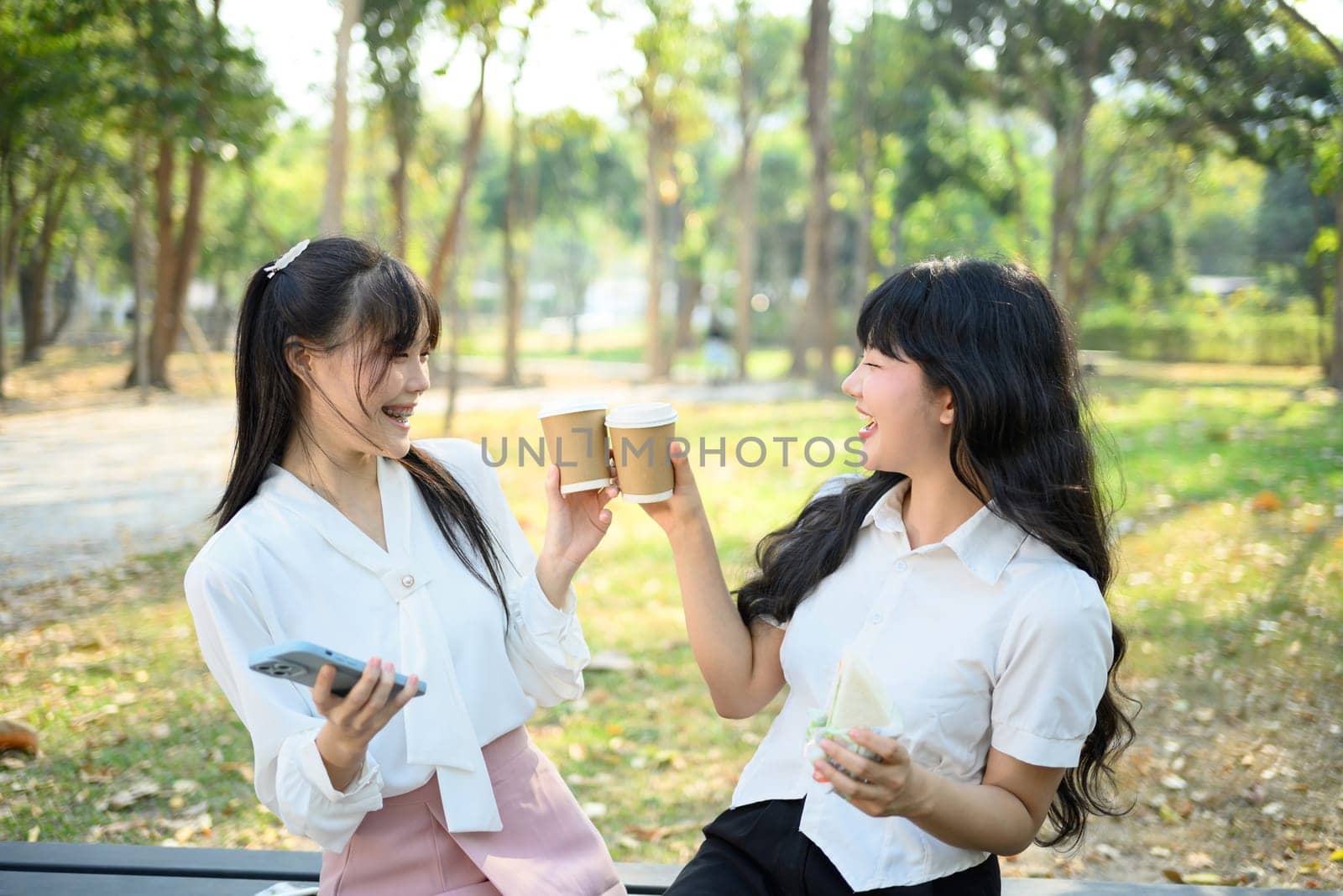 Happy young women office workers sitting on park bench and drinking coffee by prathanchorruangsak