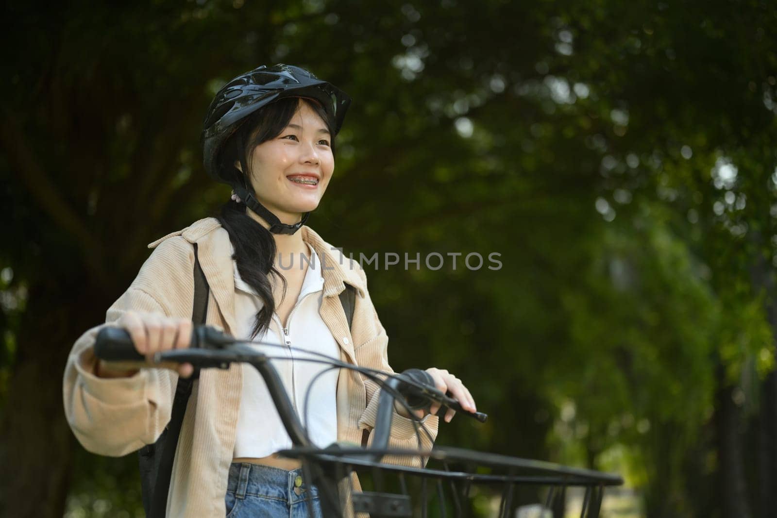 Smiling young woman wearing helmet walking in the park with her bicycle.