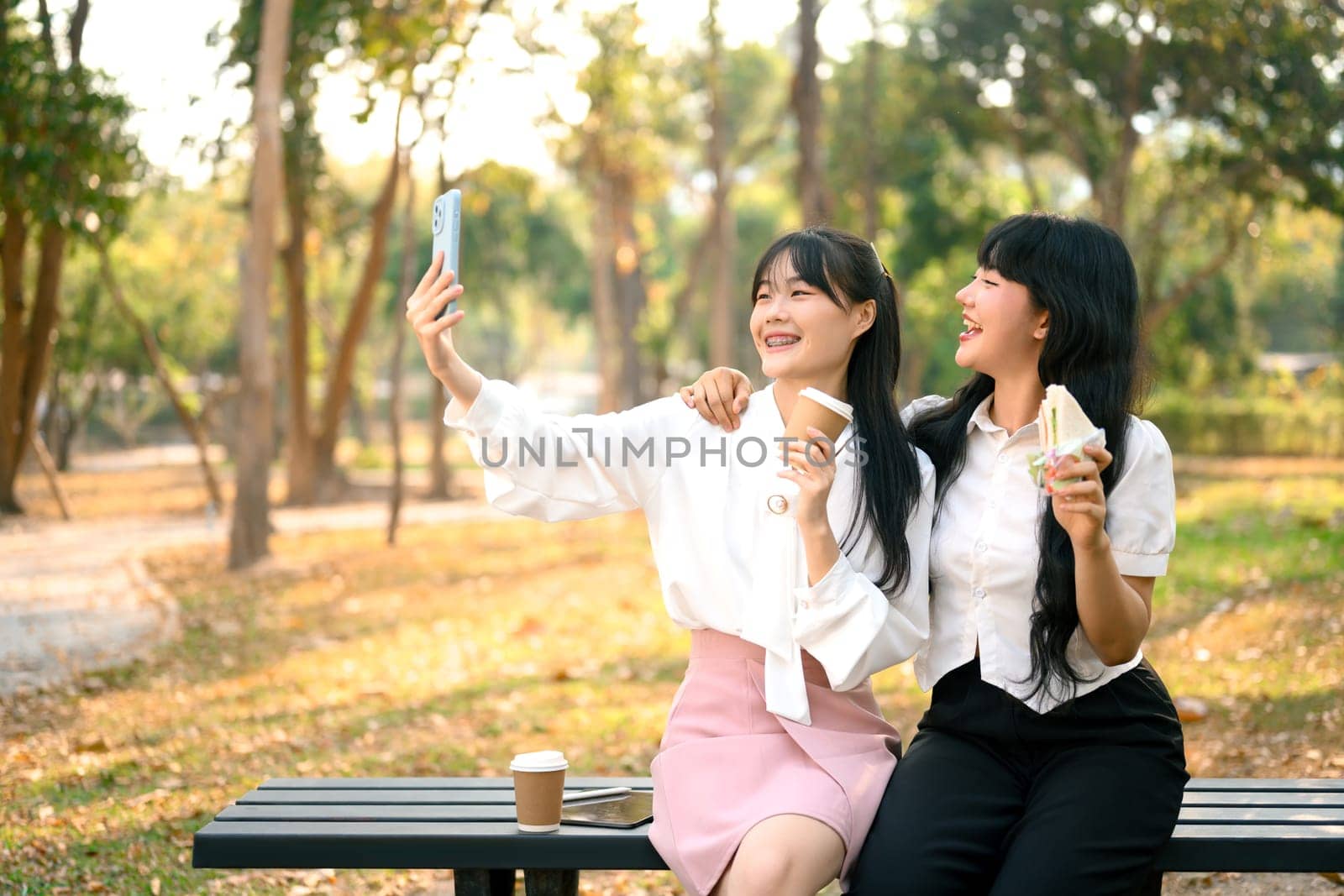 Two woman friends sitting on bench in public park and taking a selfie with mobile phone by prathanchorruangsak