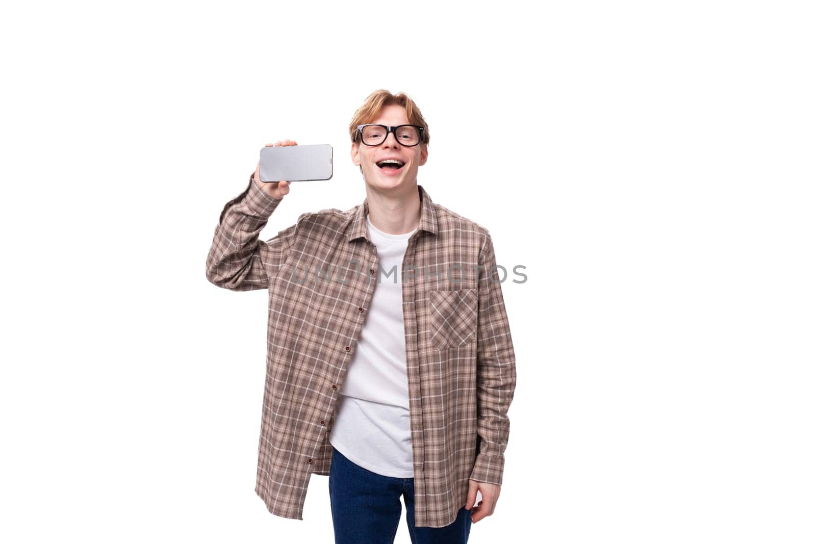 young caucasian guy with red hair in glasses and a plaid shirt shows the screen of a mobile phone with a mockup.