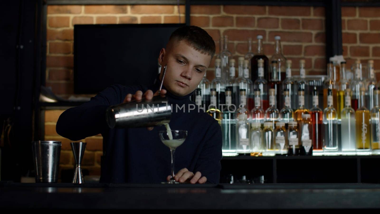 The barman preparing perfect cocktail, standing behind the bar counter. Media. Many bottles of alcohol on the background. by Mediawhalestock