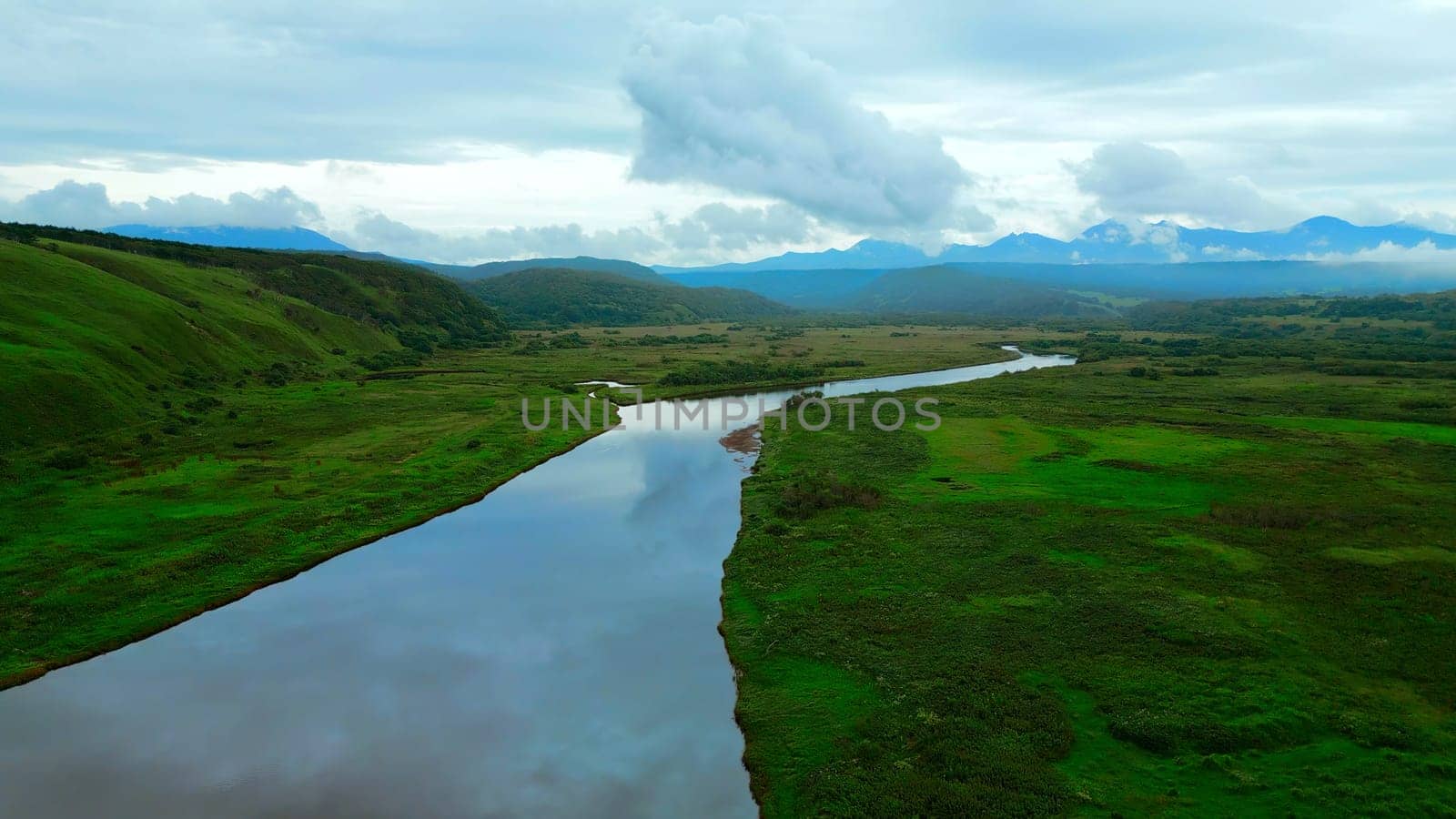 Flying above river and green valley as a backdrop. Clip. Aerial view of summer green meadow and long narrow river