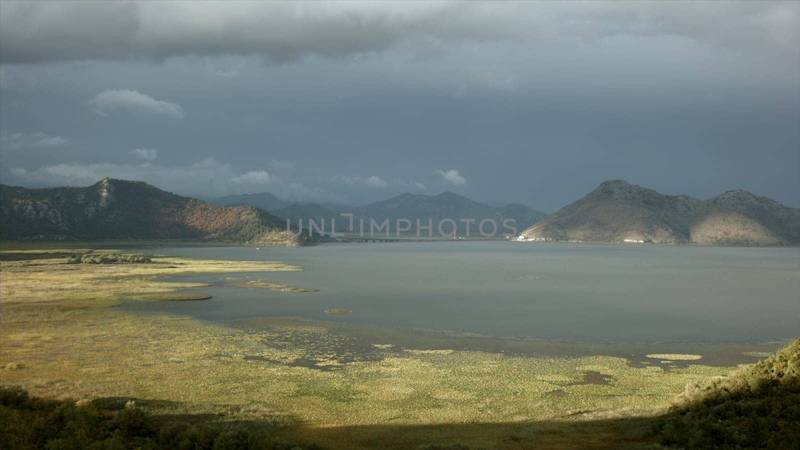 Sun and shadow on green meadow and a lake. Creative. Calm water and mountains on cloudy sky background. by Mediawhalestock