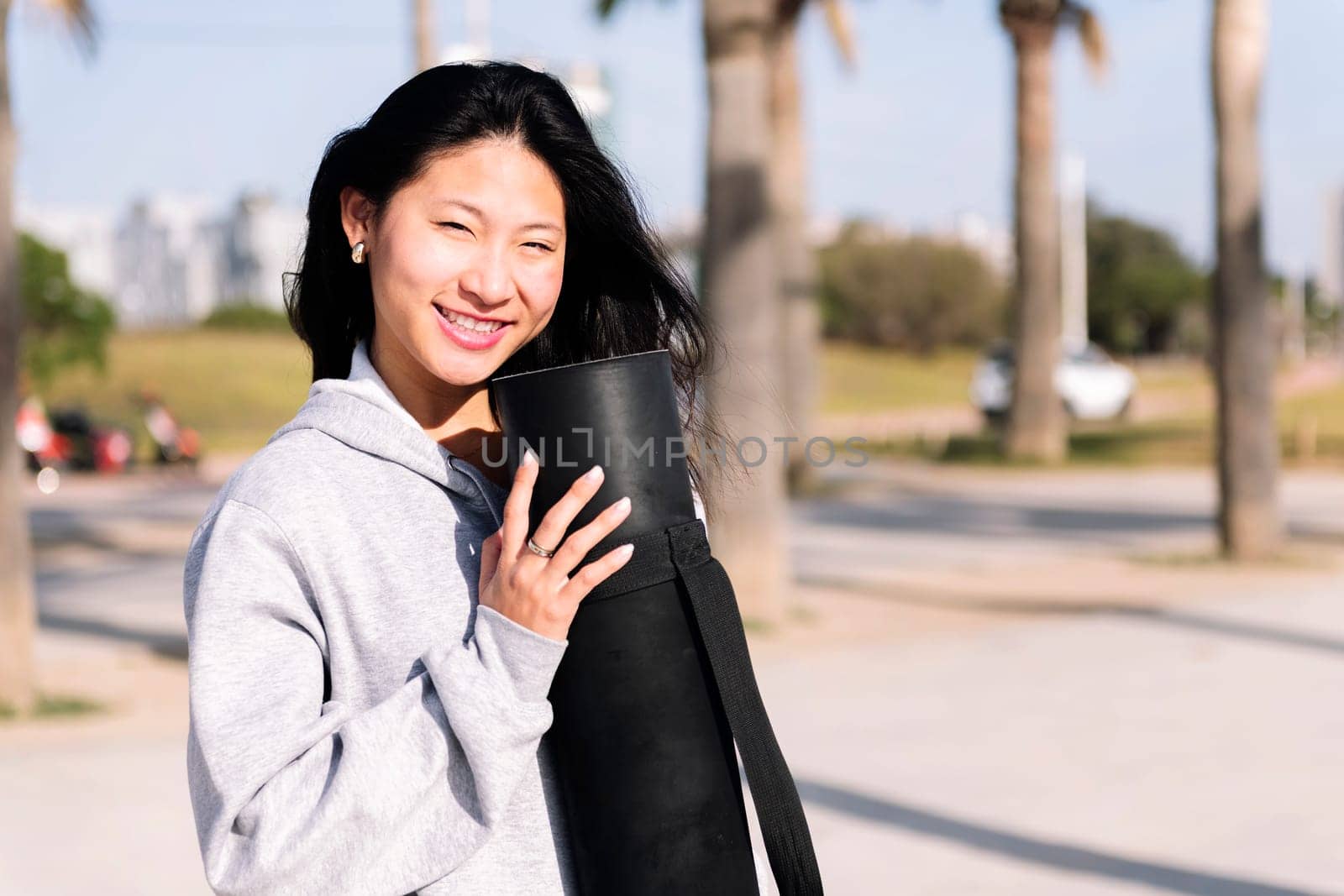 portrait of a young asian woman dressed in casual clothes smiling looking at camera while holding her yoga mat, healthy and active lifestyle concept, copy space for text
