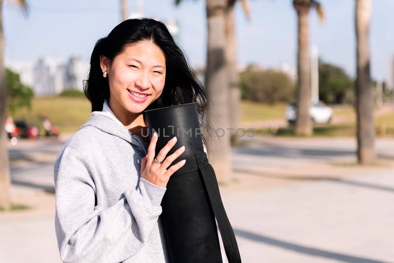 portrait of a young asian woman dressed in casual clothes smiling looking at camera while holding her yoga mat, healthy and active lifestyle concept, copy space for text