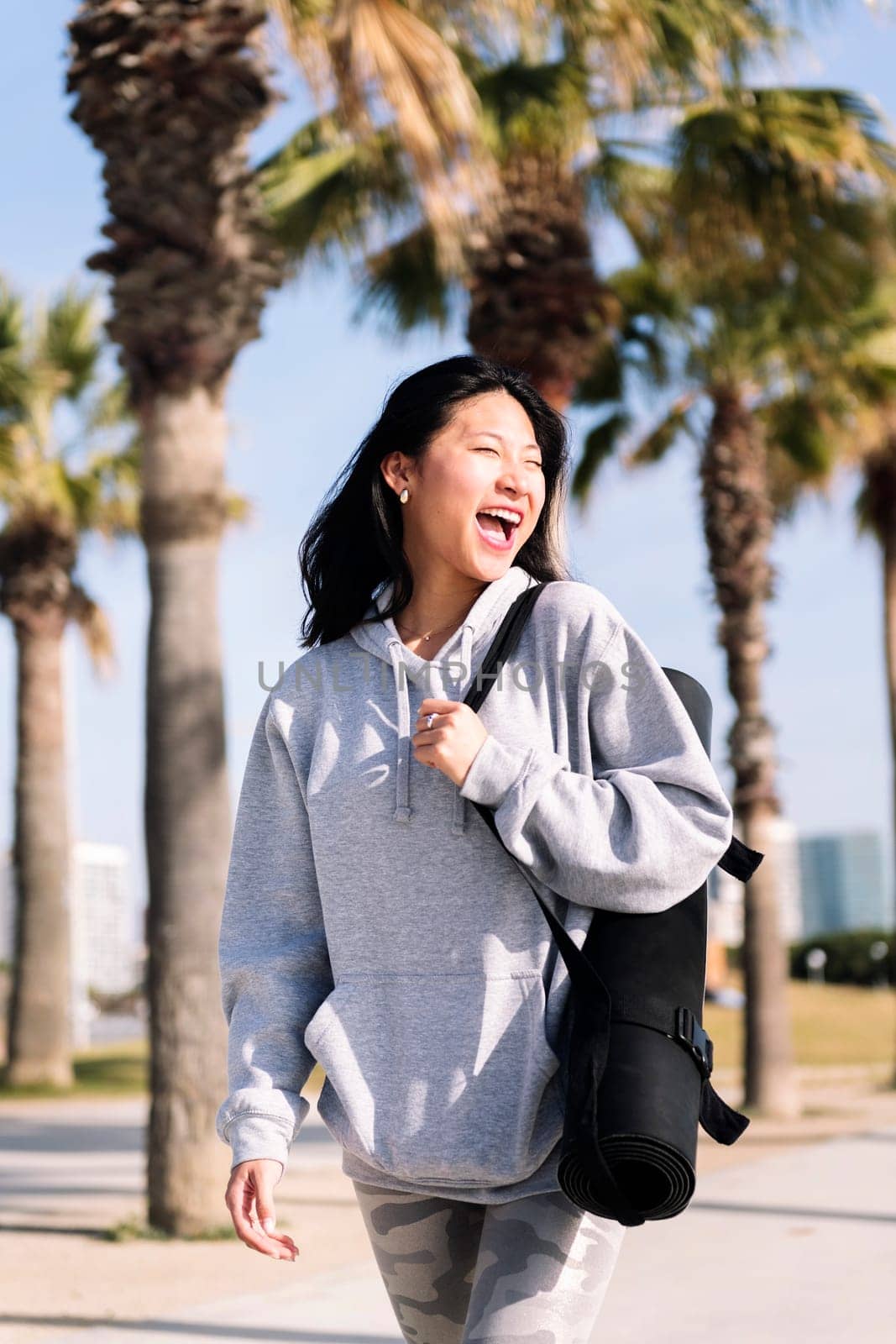young asian woman dressed in sportswear laughing happy while walking with yoga mat hanging on her arm, sport and healthy lifestyle concept