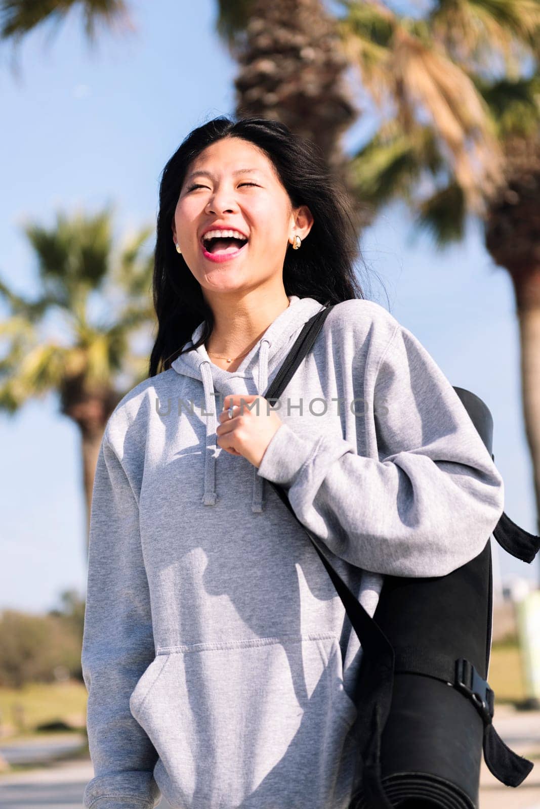 young asian woman dressed in sportswear laughing happy while walking with yoga mat hanging on her arm, sport and healthy lifestyle concept