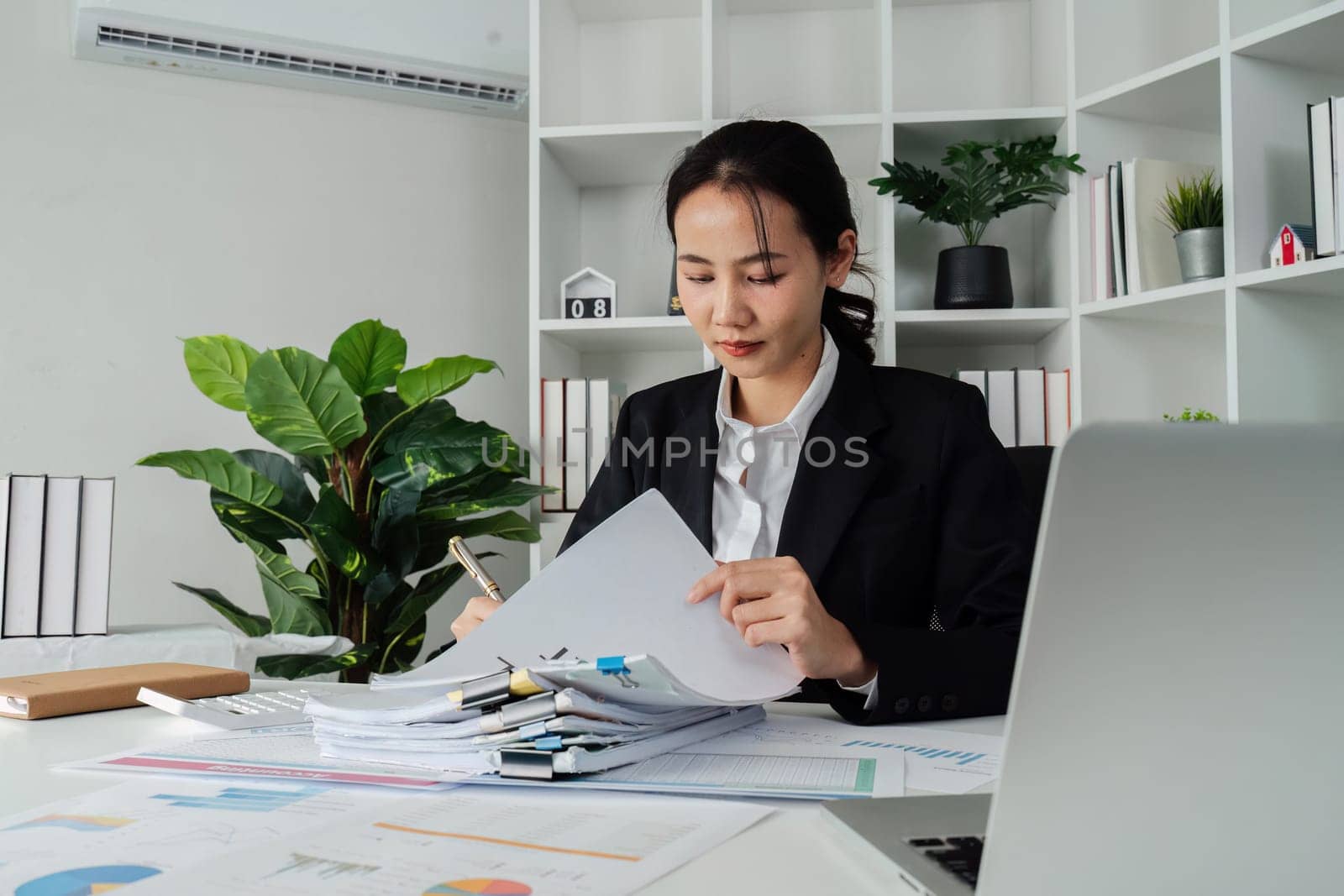 A woman is writing on a piece of paper with a pen. She is wearing a black suit and is sitting at a desk. The paper has a lot of numbers and calculations on it