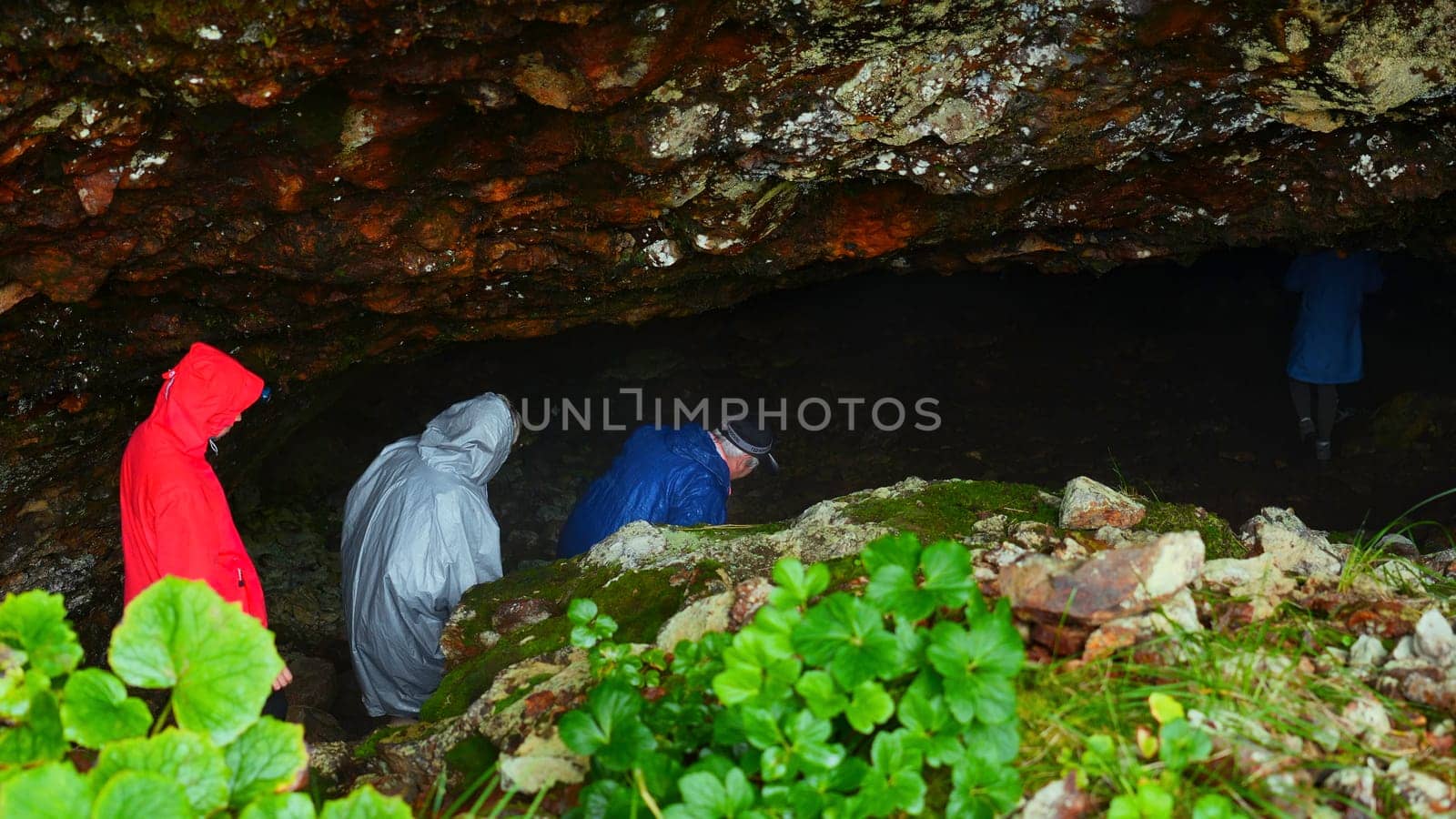 Group of tourists go to cave. Clip. Tourists enter cave in rocks on rainy day. Group of people in raincoats in rocky Mountains go to cave in summer.