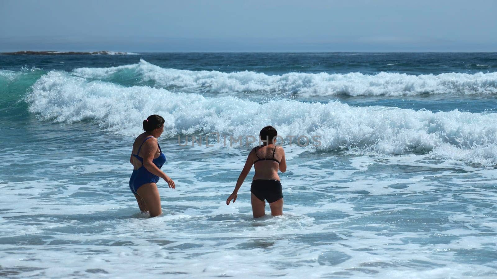 People bathe in beautiful blue waves on sunny day. Clip. Beautiful blue waves with foam on shore with vacationing people. People in swimsuits swim with blue waves of sea on summer day.