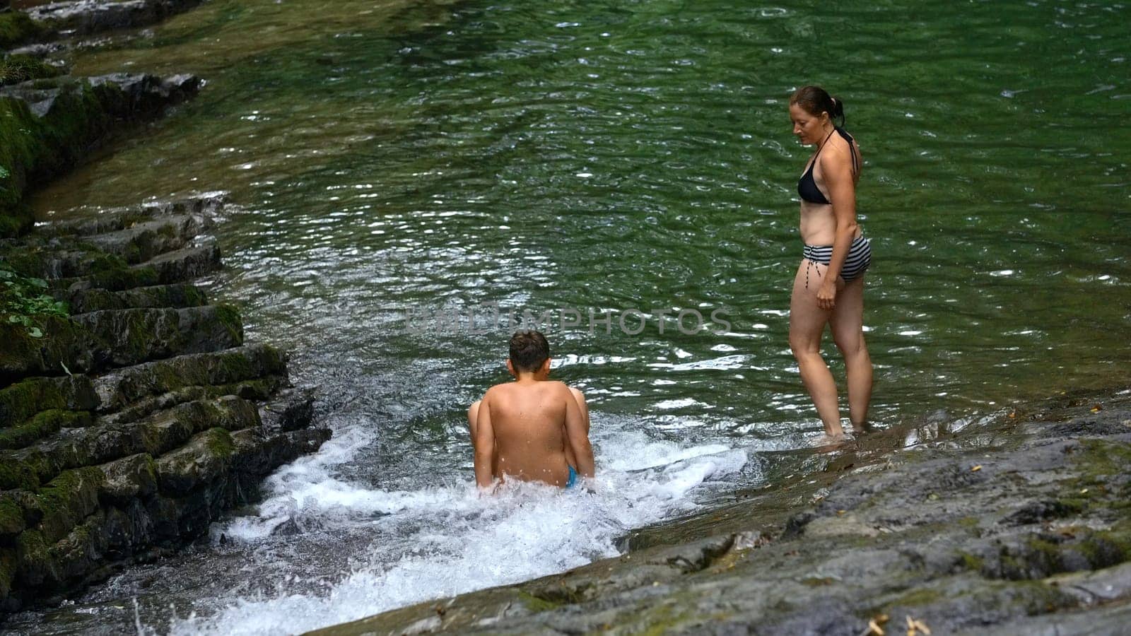 Woman and teenage boy bathing in splashing waterfall in tropical forest. Creative. Happy family enjoying fresh water from flowing waterfall in jungle