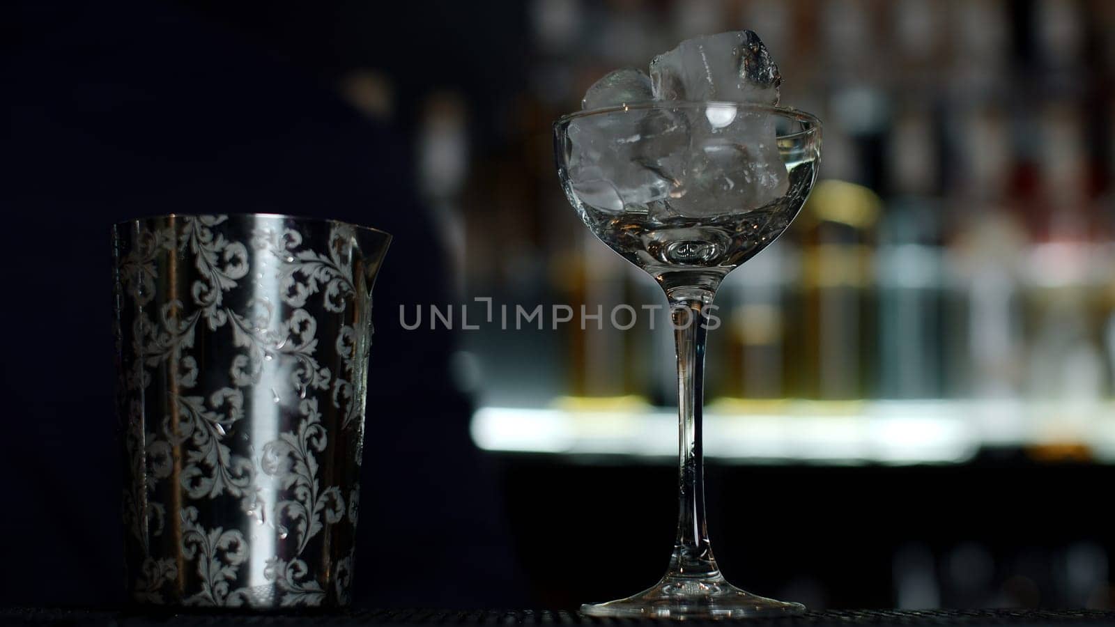 Close up of a bartender putting ice cubes into the small empty cocktail glass. Media. Details of making a drink with blurred bottles of alcohol on the background