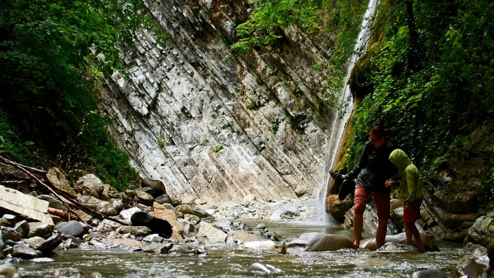 Mother and boy crossing mountain cold water stream with stones. Creative. Hikers in picturesque place. by Mediawhalestock