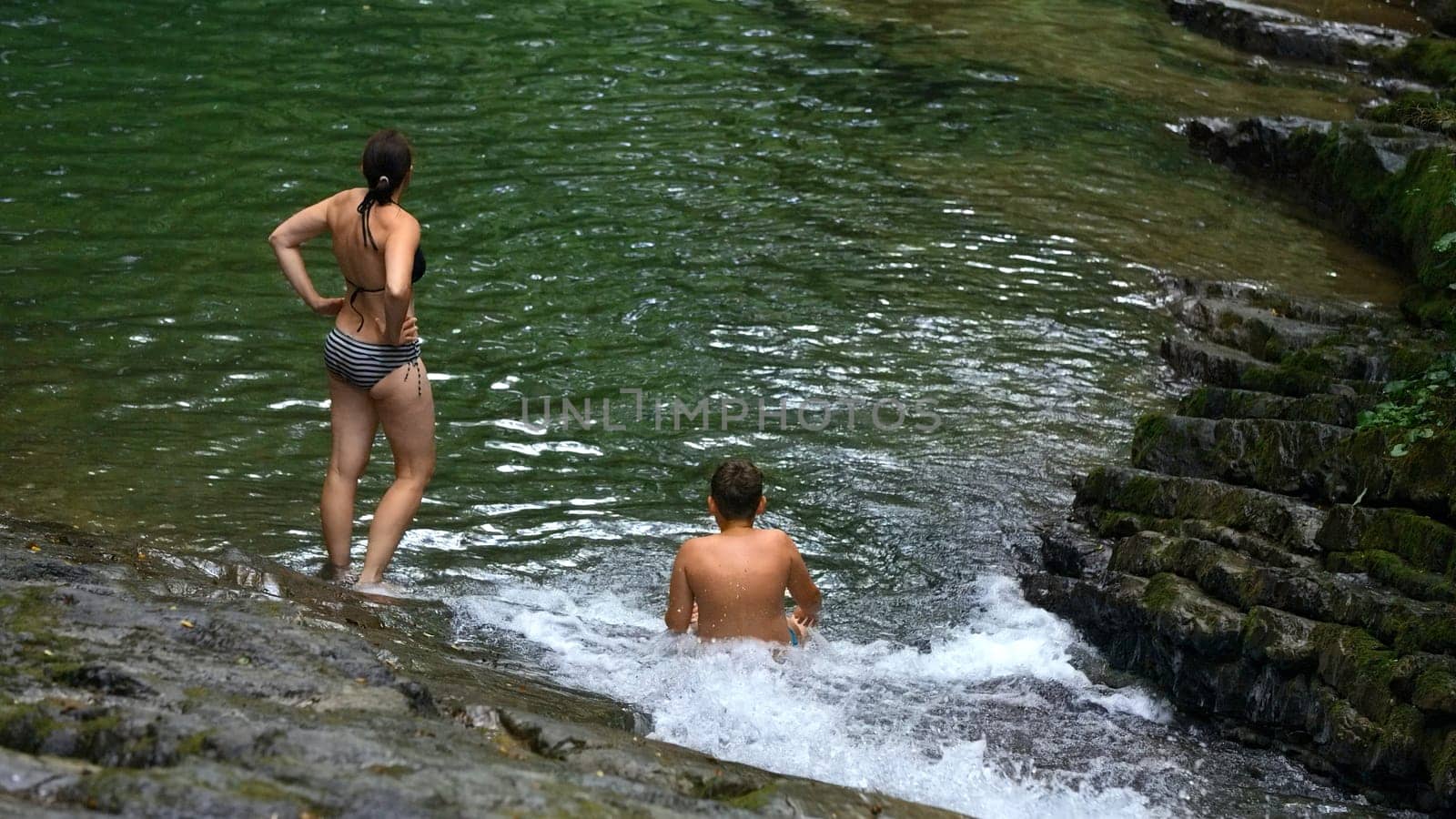 Woman and teenage boy bathing in splashing waterfall in tropical forest. Creative. Happy family enjoying fresh water from flowing waterfall in jungle. by Mediawhalestock