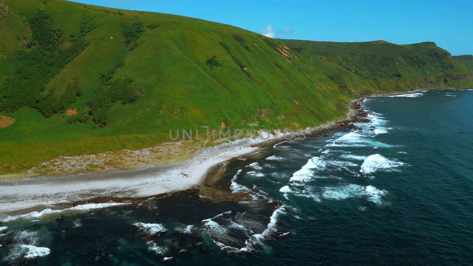 Wild nature, green mountains with steep green slopes leading to the sea. Clip. Aerial of foam of blue waves break in volcanic rocks