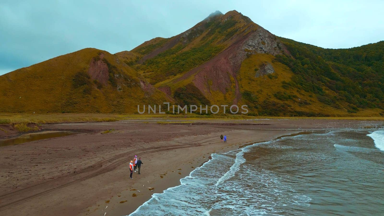 Top view of people by sea with mountain view. Clip. Coast with walking people and amazing mountains on cloudy day. Walk on mountain coast of ocean on cloudy day.