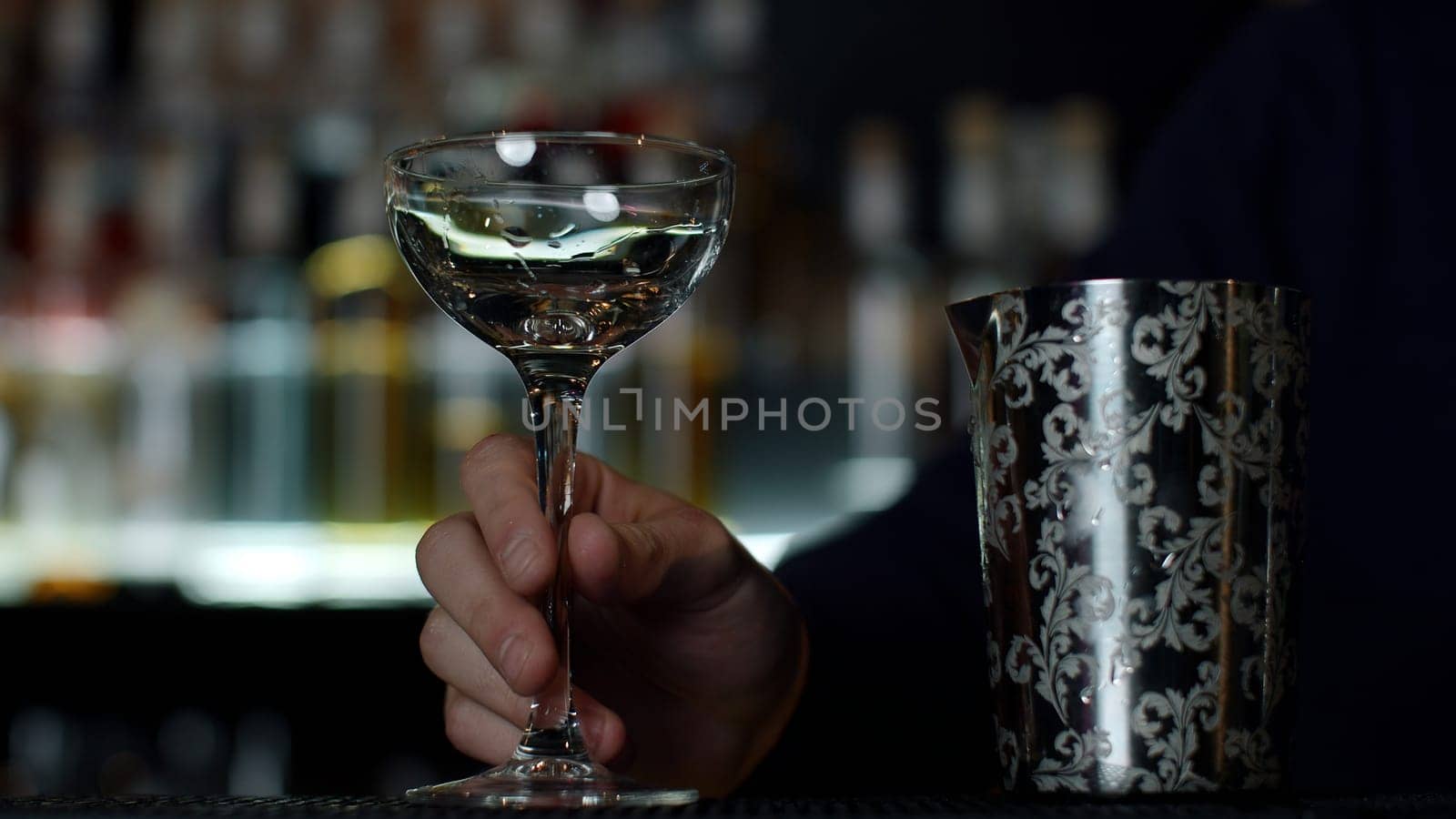 Close up of a bartender putting ice cubes into the small empty cocktail glass. Media. Details of making a drink with blurred bottles of alcohol on the background. by Mediawhalestock