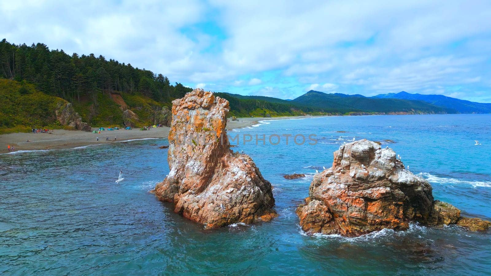 Top view of rocks in sea and coast with tourists. Clip. Beautiful coast with rocks and turquoise water. Camping vacation on wild coast of sea with rocks by Mediawhalestock