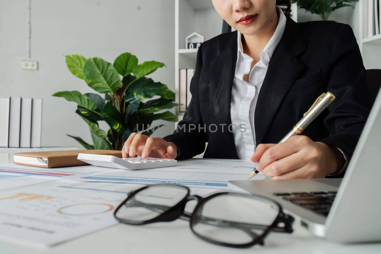 A woman is writing on a piece of paper with a pen. She is wearing a black suit and is sitting at a desk. The paper has a lot of numbers and calculations on it