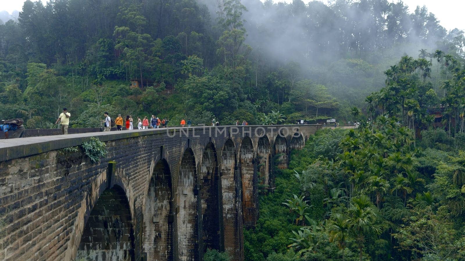 Ancient stone aqueduct in green mountains. Action. Ancient stone bridge with arches in green mountains. Tourists walk on ancient bridge in valley of tropical mountain forests.