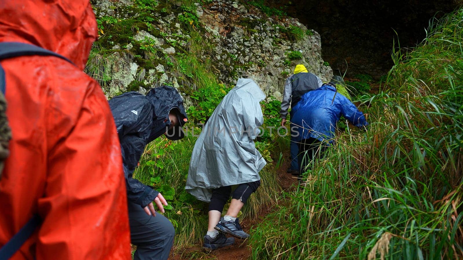 Group of tourists go to cave. Clip. Tourists enter cave in rocks on rainy day. Group of people in raincoats in rocky Mountains go to cave in summer by Mediawhalestock