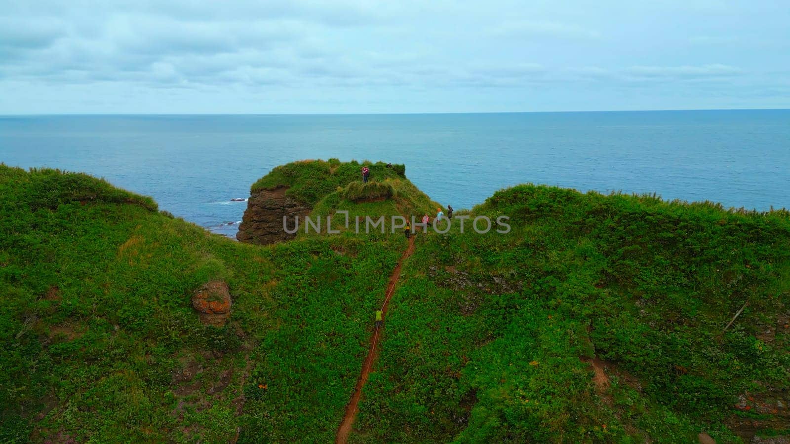 Top view of group of people standing on edge of coast. Clip. Tourists are standing on rocky shore overlooking sea horizon. Tourists on rock with beautiful grass by sea on cloudy day by Mediawhalestock