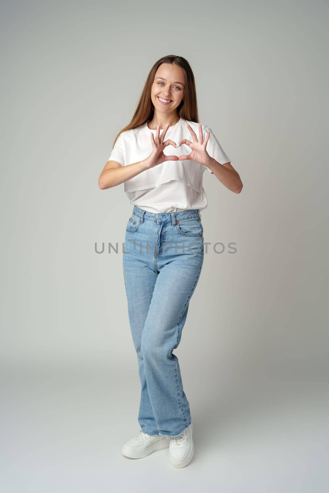 Smiling young woman showing heart sign with hands on gray background by Fabrikasimf