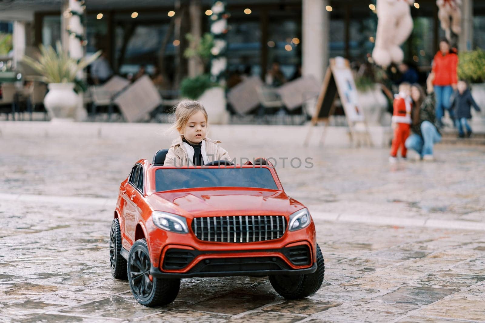 Little girl drives a red toy car across the square. High quality photo