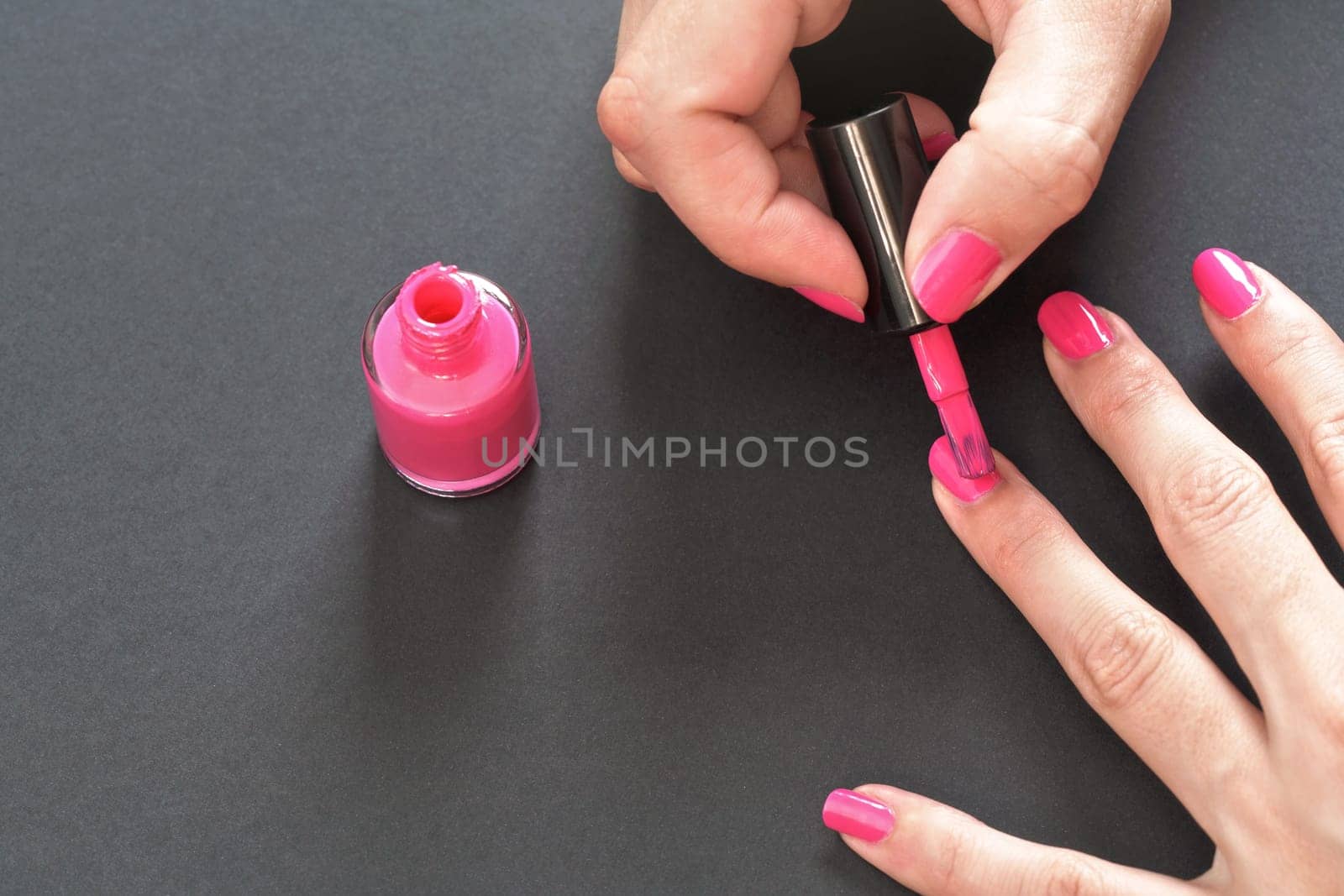 Applying pink nail polish - closeup photo of woman hands, little brush over fingernails, small bottle on gray / black background