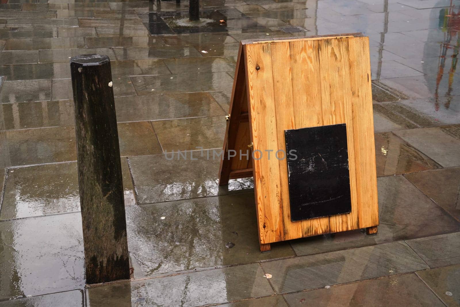 Wooden stand with small empty black board (space for your text) on street wet from rain