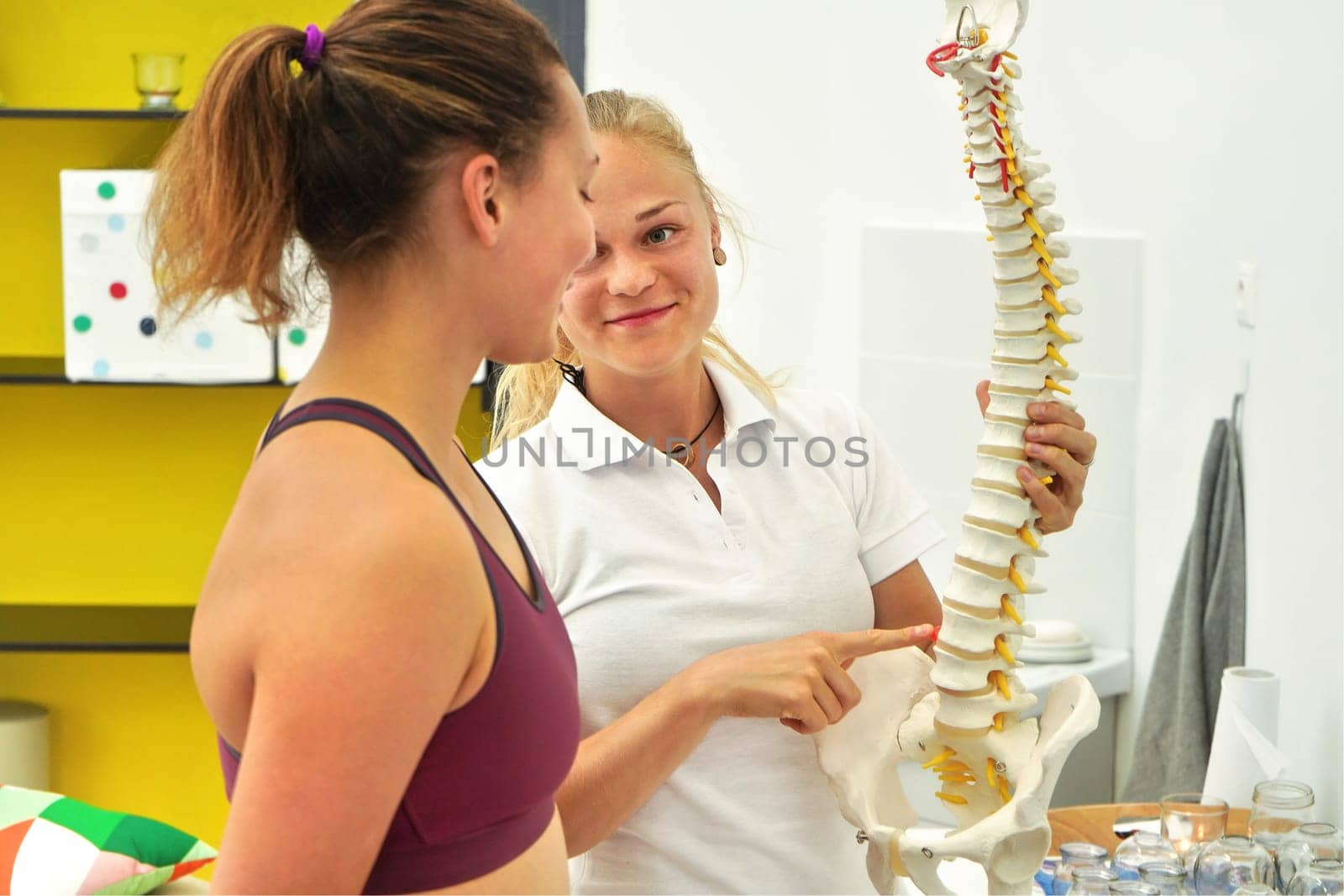 Young woman patient at physiotherapy consultation, physiotherapist explaining back bones on spine model by Ivanko