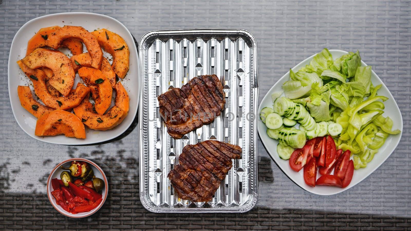 Grilled butternut squash and pork cuts, some fresh vegetable next to it, on glass garden table, view from above